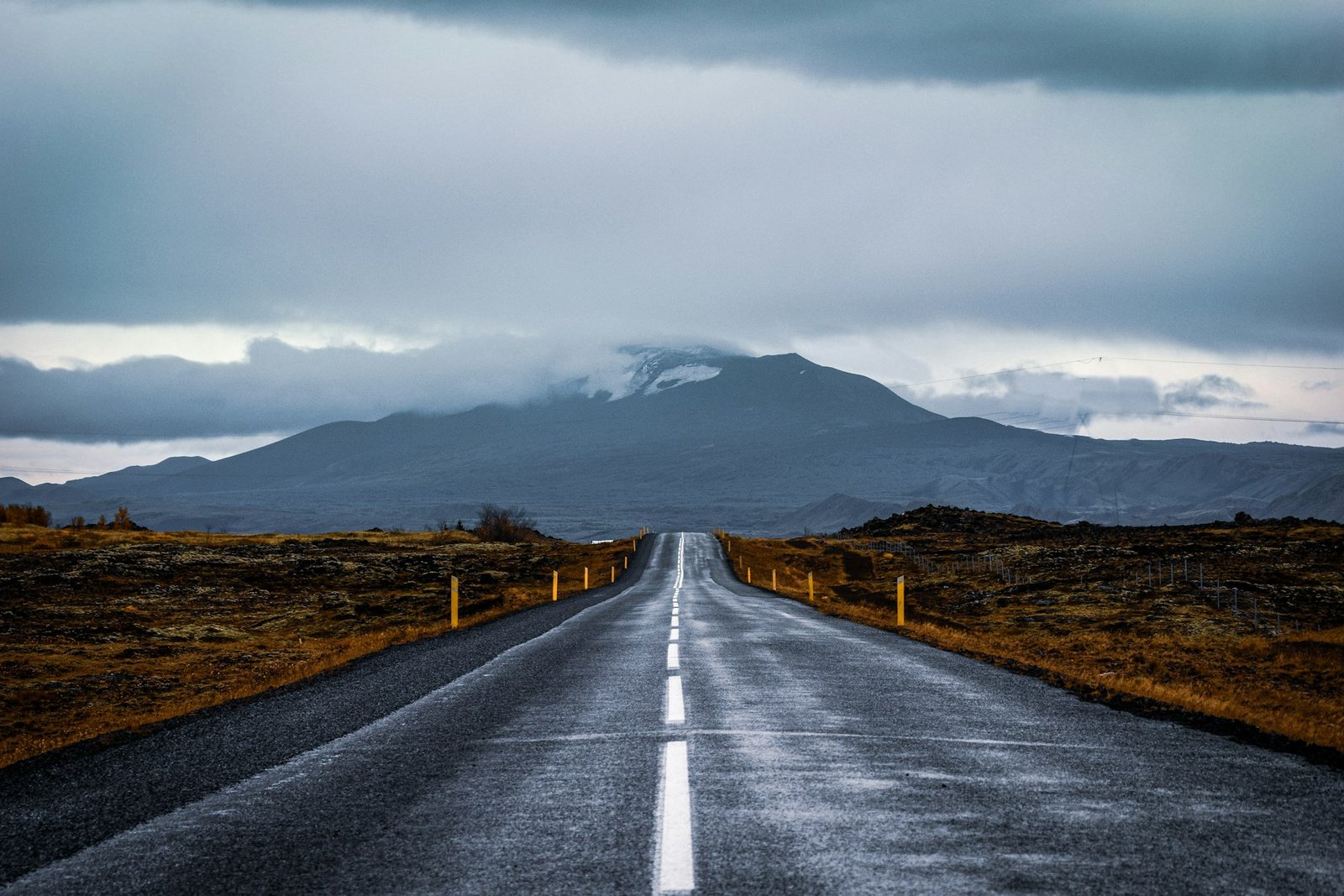 A grey road running towards a dark Icelandic mountain under a white sky